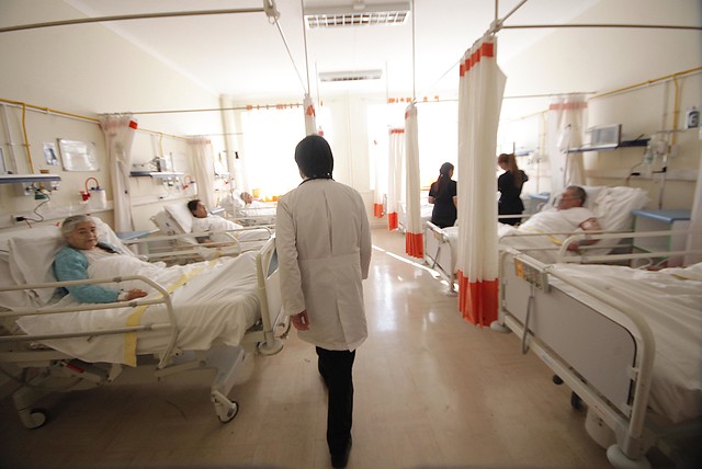 La imagen muestra a una sala de hospital con varios pacientes y un doctor caminando de espaldas