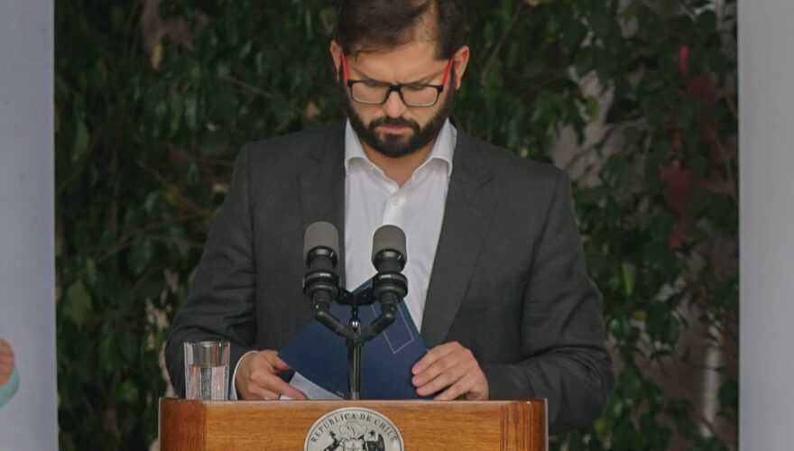 Gabriel Boric frente a un podio con el logo presidencial mirando hacia abajo como revisando unas carpetas que toma con sus manos