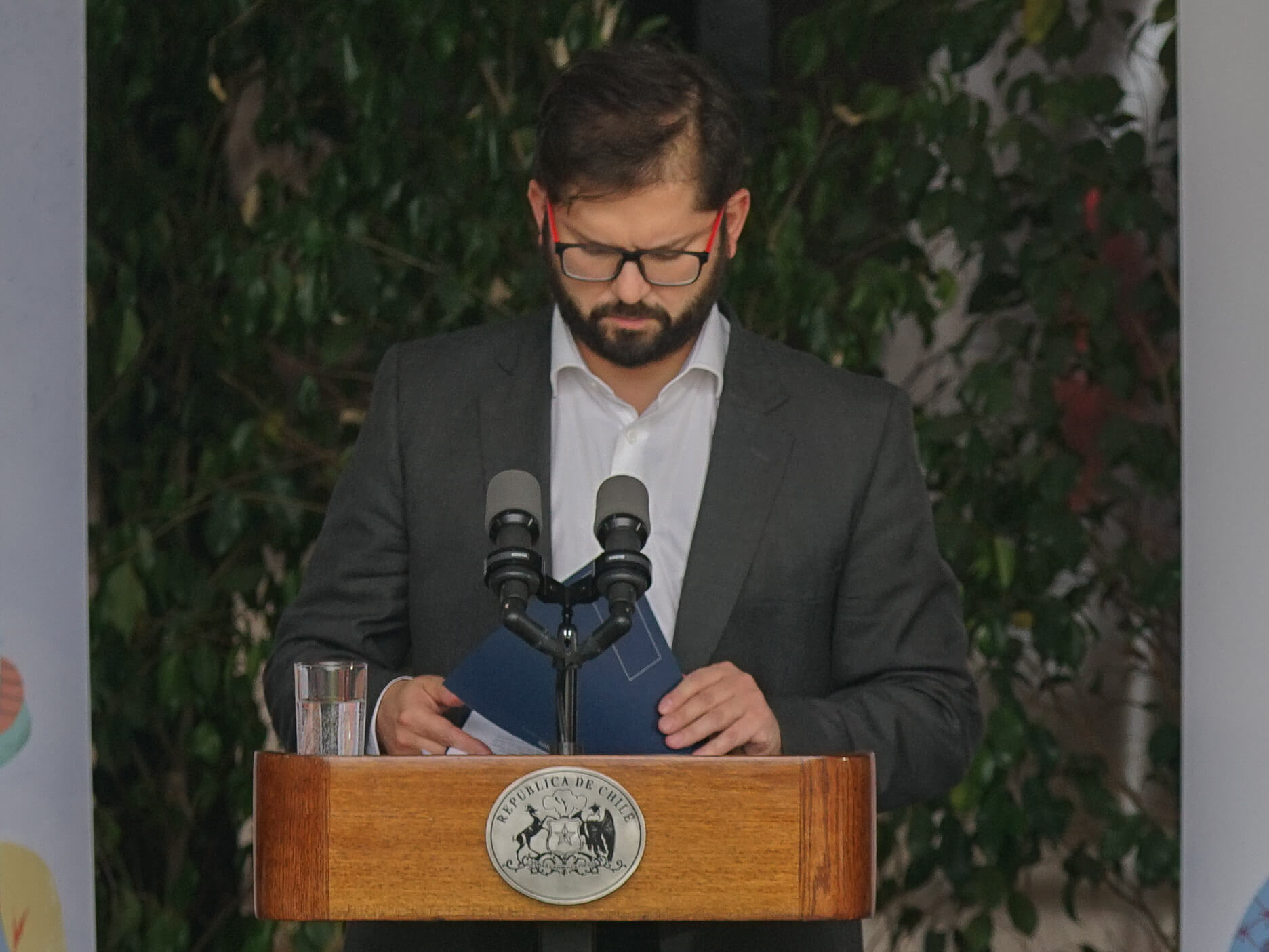 Gabriel Boric frente a un podio con el logo presidencial mirando hacia abajo como revisando unas carpetas que toma con sus manos